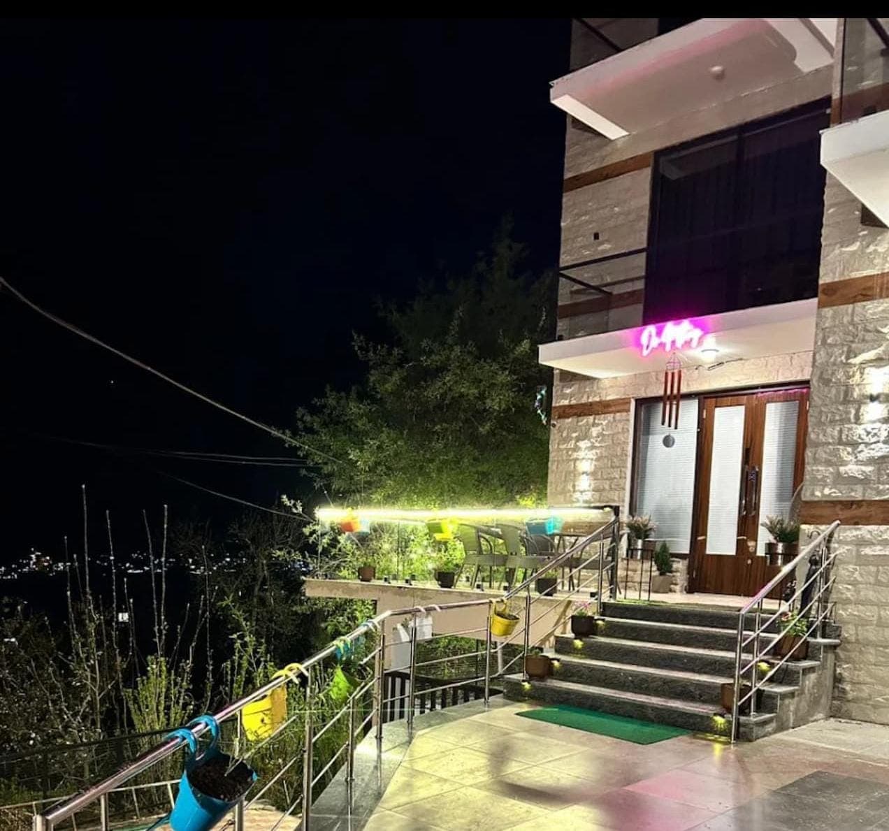 Cafe DO NTHNG at night, its neon sign glowing above a stone entrance with terrace seating and potted plants