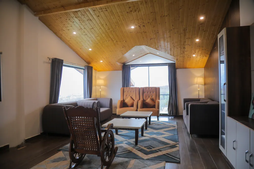 Lounge under a pitched timber ceiling with sofas, orange armchairs, a rocking chair and a window towards the valley