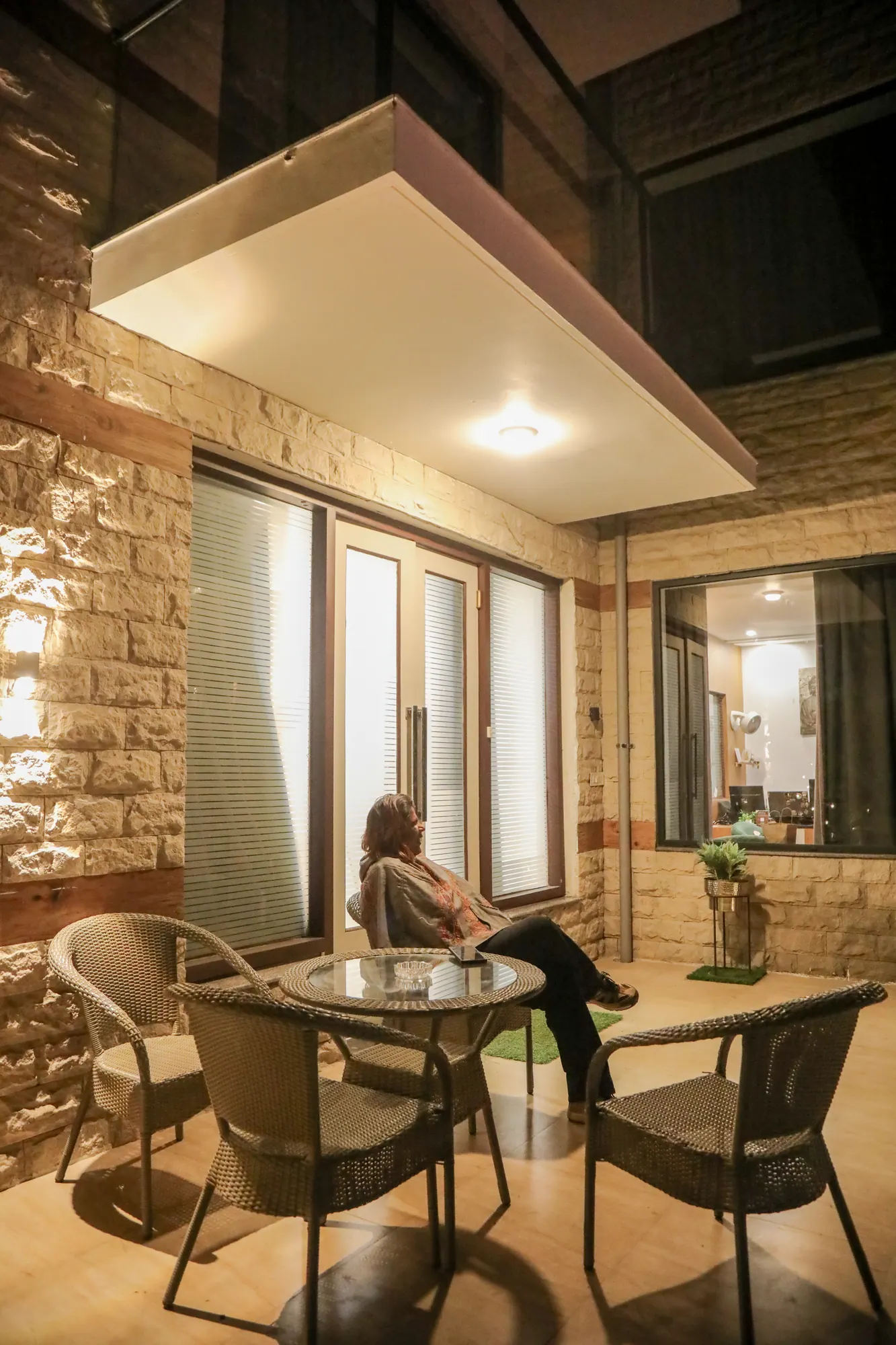 Terrace with wicker chairs and a glass table beside stone-clad walls and glass doors, with a guest seated at the edge