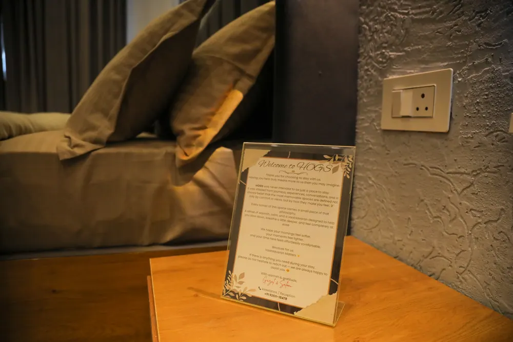 Framed welcome card on a wooden bedside table beside a bed with cushions and a textured grey wall