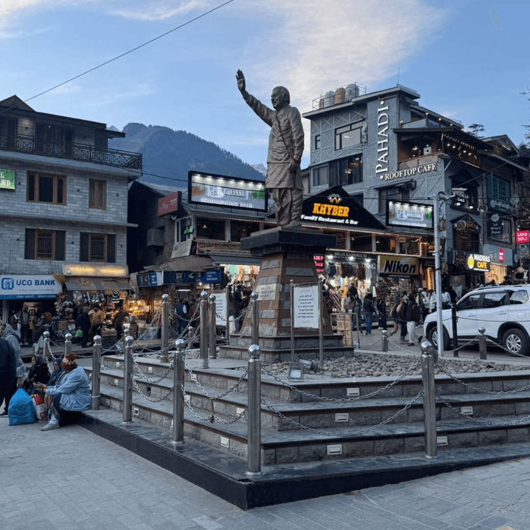 Mall Road in central Manali at dusk, with a bronze statue, rooftop cafes and shop signs under a blue sky