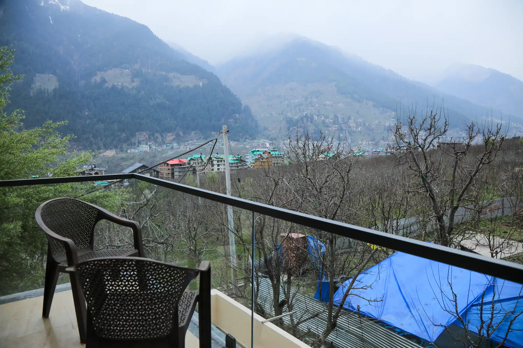 Balcony with two chairs and a glass railing overlooking bare orchard trees, rooftops and misty mountains at HOGS Panorama, Manali