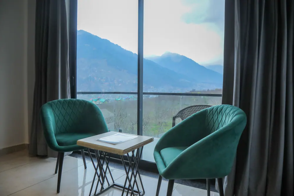 Two green armchairs and a small table beside a floor-to-ceiling window framing a mountain ridge